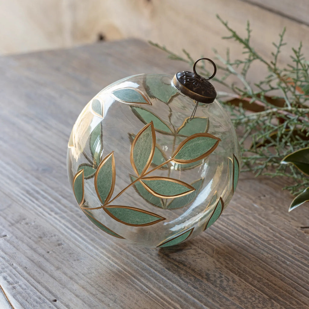 A clear glass Christmas ornament decorated with hand-painted sage green leaves and gold outlines, resting on a rustic wooden surface next to greenery.