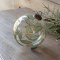 A clear glass Christmas ornament decorated with hand-painted sage green leaves and gold outlines, resting on a rustic wooden surface next to greenery.
