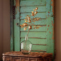 A decorative gold artificial botanical branch with metallic leaves and round gold accents, arranged in a clear glass bottle vase resting on a woven wicker surface against a distressed teal-green wooden shutter background.