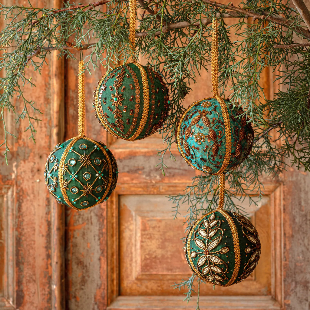 Four decorative hanging Christmas ornaments shaped like spheres, decorated with green and turquoise patterns and gold trim, hanging from a pine branch in front of a rustic wooden door.