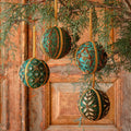 Four decorative hanging Christmas ornaments shaped like spheres, decorated with green and turquoise patterns and gold trim, hanging from a pine branch in front of a rustic wooden door.