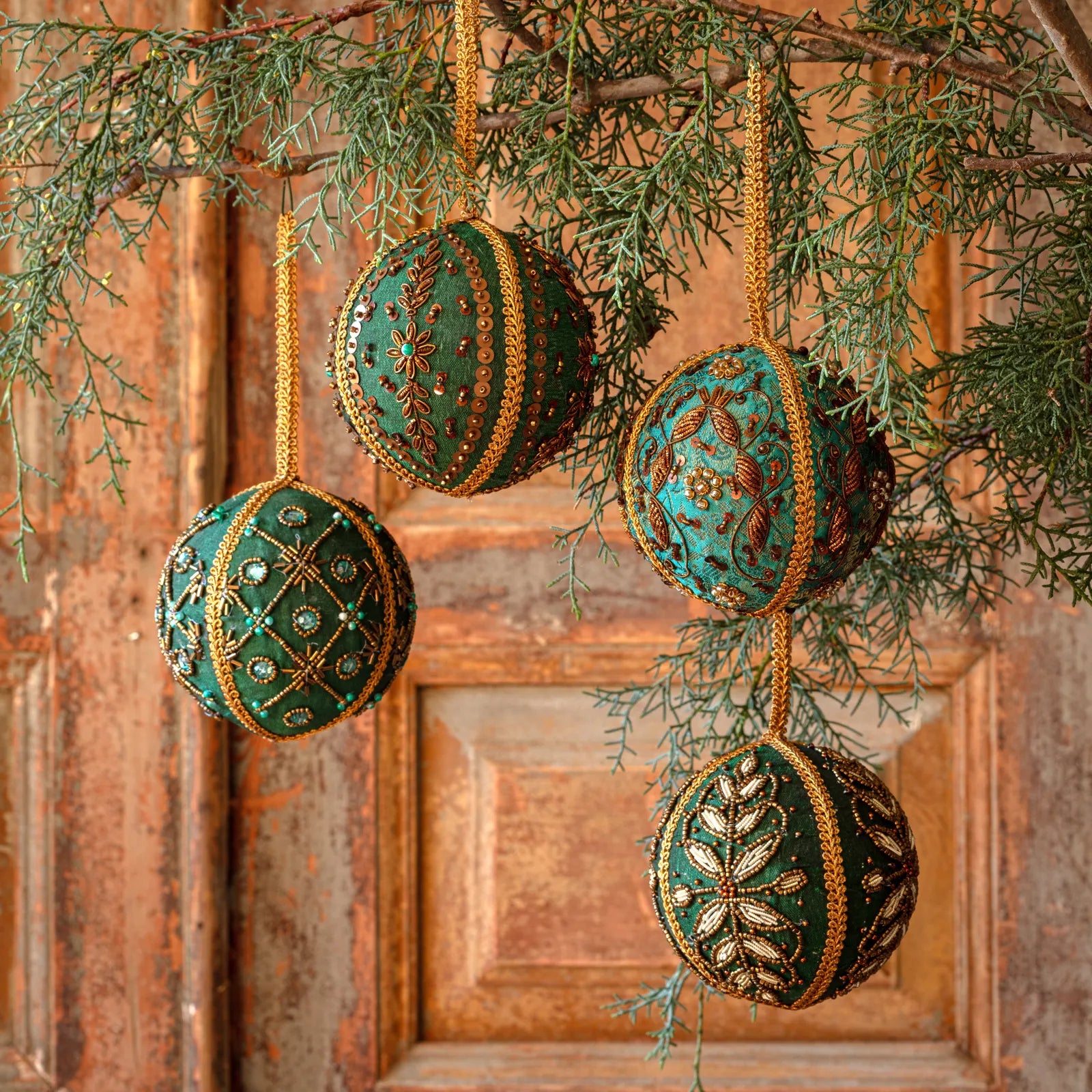 Four decorative hanging Christmas ornaments shaped like spheres, decorated with green and turquoise patterns and gold trim, hanging from a pine branch in front of a rustic wooden door.