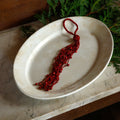 A red beaded tassel ornament with a loop at the top, displayed on an oval cream-colored ceramic dish with green foliage in the background.