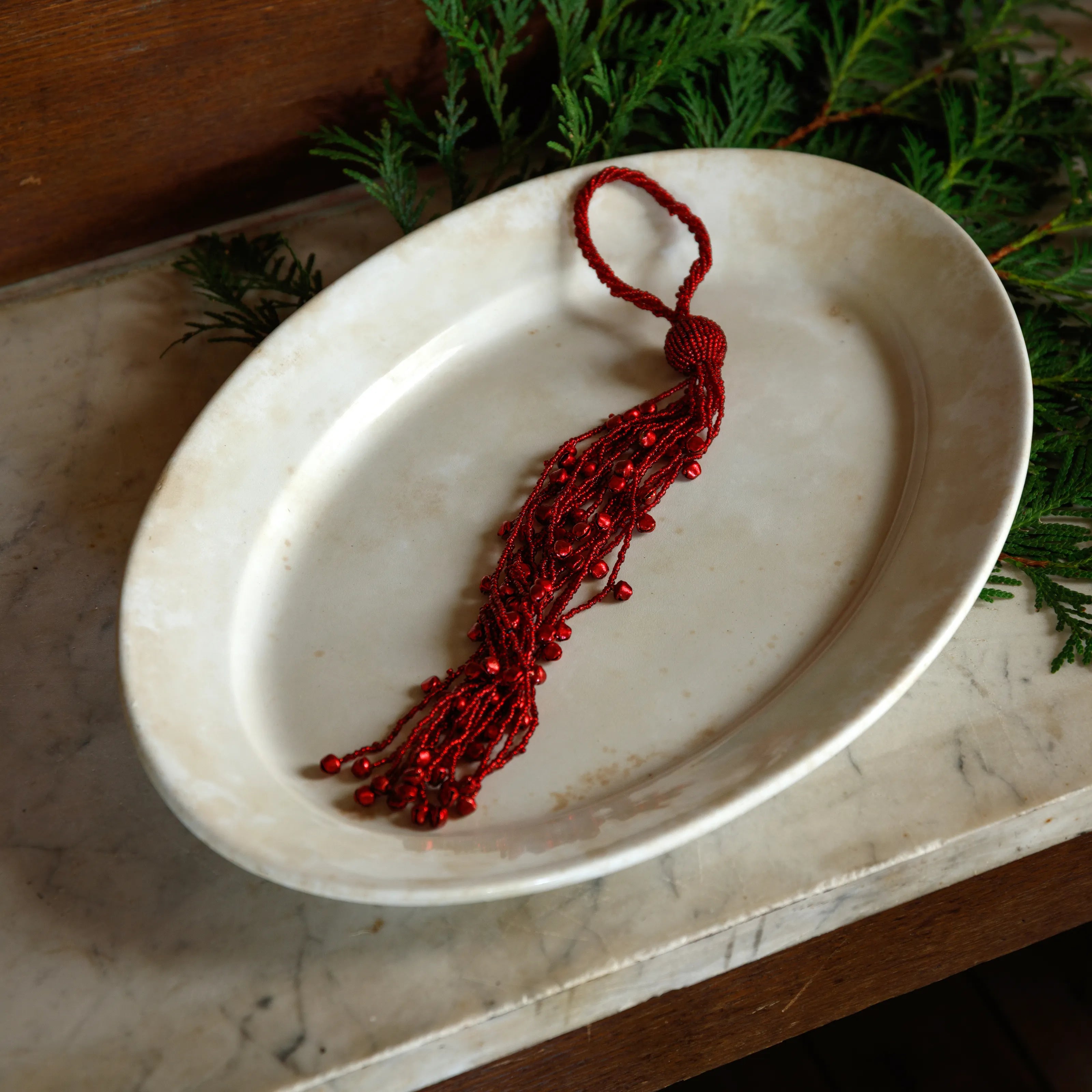 A red beaded tassel ornament with a loop at the top, displayed on an oval cream-colored ceramic dish with green foliage in the background.
