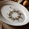 A gold-toned floral branch wreath ornament with hanging loops, displayed on a round off-white ceramic plate over a textured brown fabric surface.