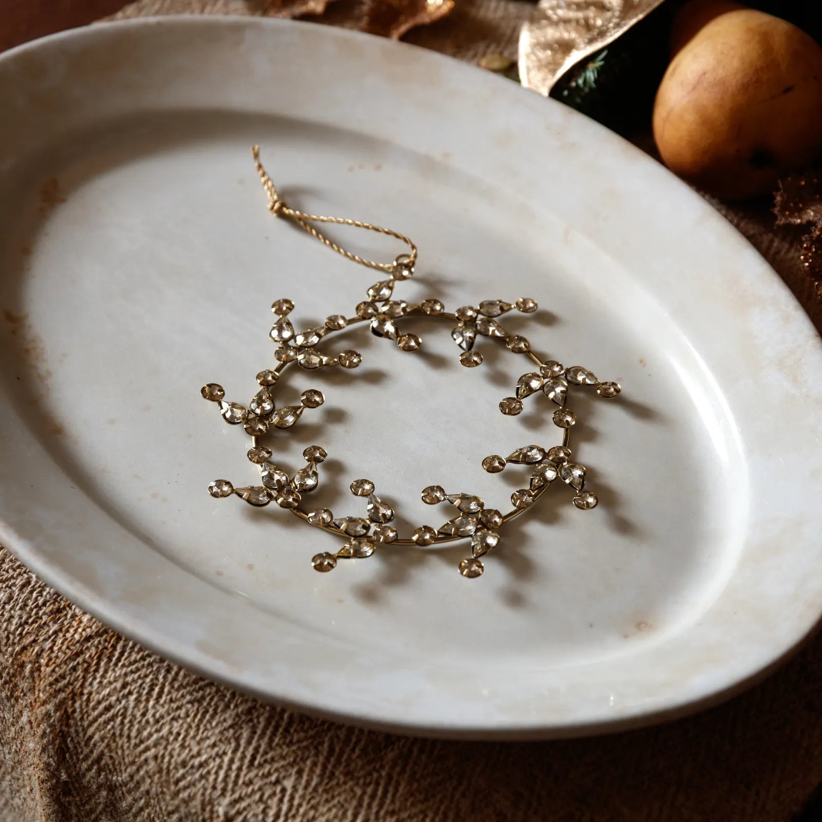 A gold-toned floral branch wreath ornament with hanging loops, displayed on a round off-white ceramic plate over a textured brown fabric surface.