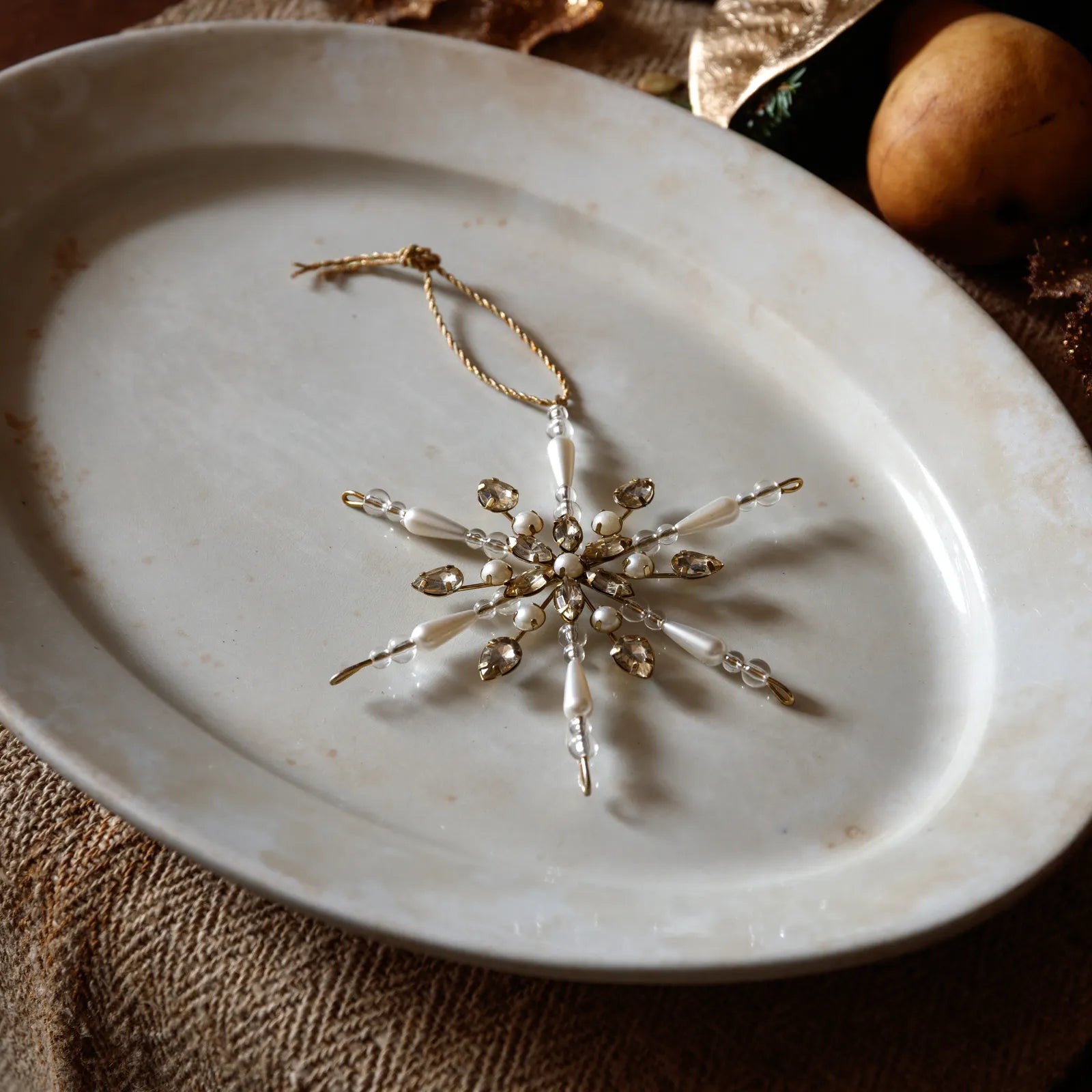 A gold and white beaded snowflake Christmas ornament with a hanging loop, displayed on a rustic white ceramic plate.