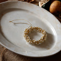A gold wreath-shaped ornament decorated with small white pearls and hanging from a thin gold string, displayed on a round off-white ceramic plate.