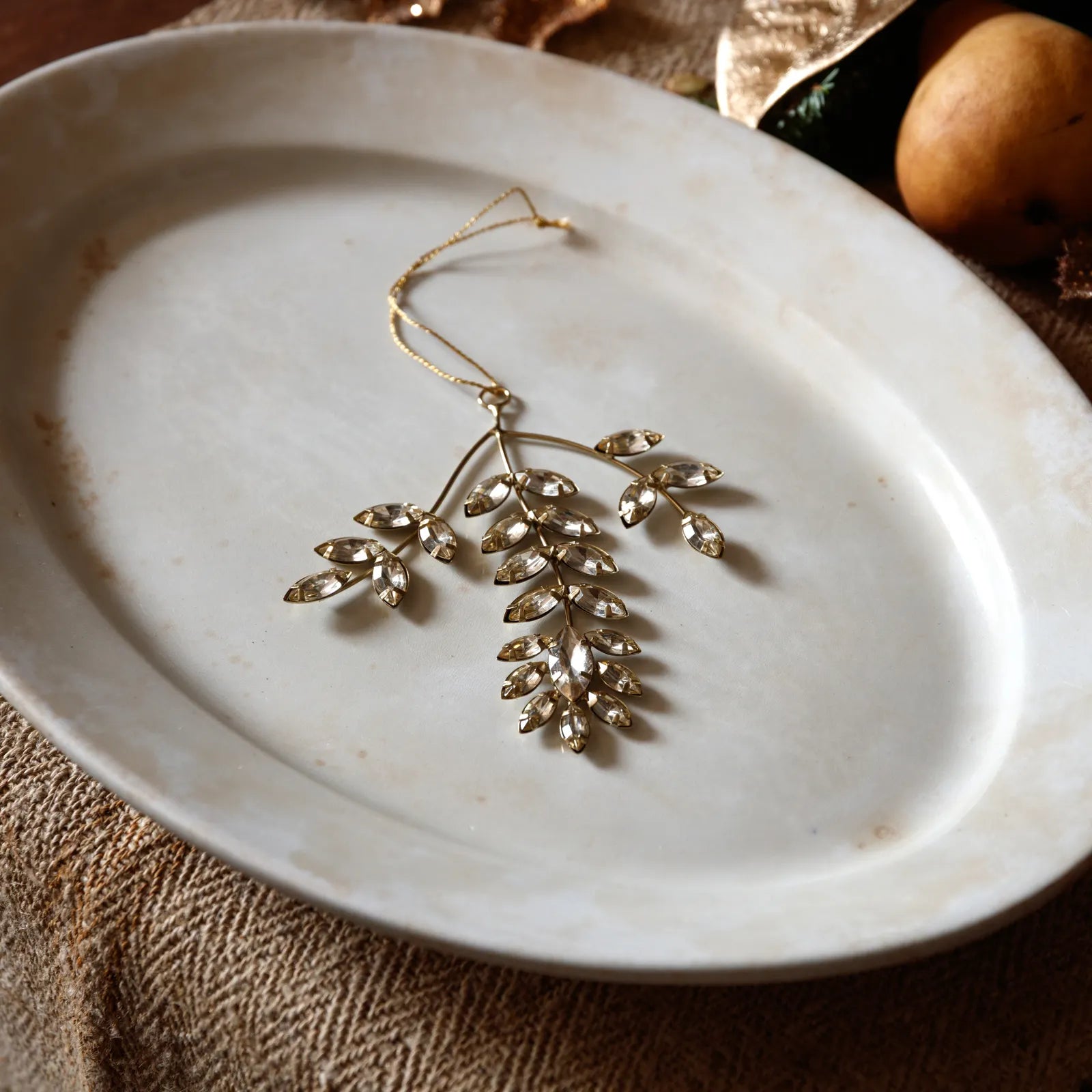 A gold-toned leaf-shaped hanging ornament embellished with clear rhinestones, resting on a round off-white ceramic plate over a textured brown fabric surface.