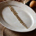A gold metallic leafy branch ornament with hanging teardrop charms, draped diagonally across a round off-white ceramic plate on a textured brown fabric surface.